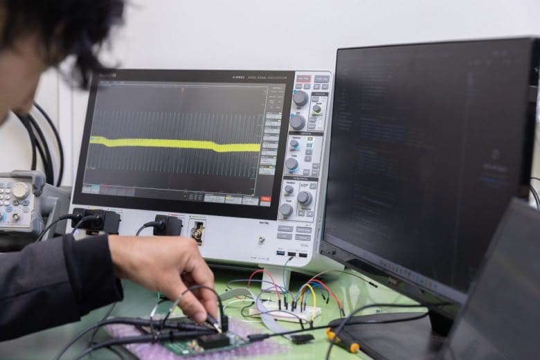 A hand touching a probe to a board while watching an oscilloscope waveform